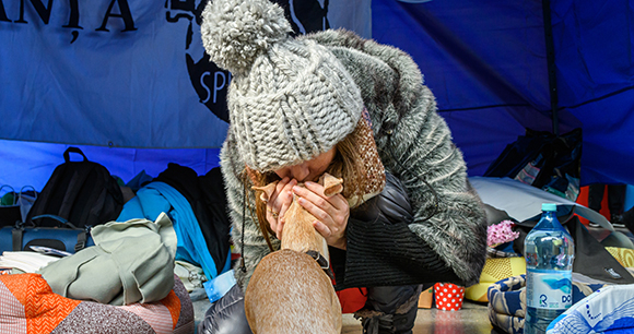 A woman crouches down while kissing her small tan dog on the head.