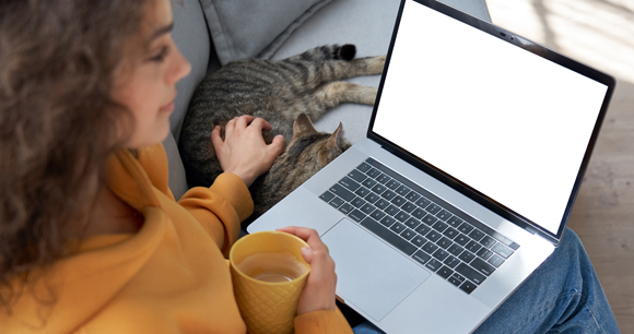 A girl with brown hair looks at her laptop screen while petting a cat.